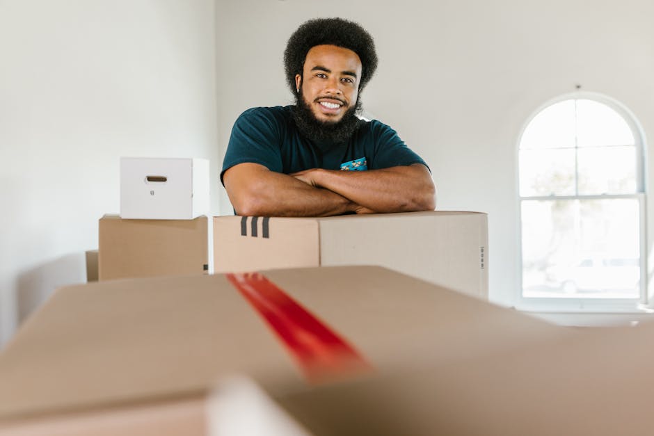 A male professional mover with a dark beard and curly hair, smiling and standing behind a stack of cardboard boxes in a well-lit room with a large arched window in the background. The boxes are of various sizes, some sealed with red packing tape, and are likely used for packing office furniture, equipment, or personal belongings as part of a house or office relocation service. The movers are inside a residential or commercial property during the packing and moving process, with the boxes placed on a clean floor near the doorway. The natural lighting coming through the window illuminates the space, highlighting the orderly arrangement of packing materials and the environment prepared for furniture transport. This scene exemplifies efficient packing and loading activities conducted by [COMPANY_NAME], engaged in house or office removals, ensuring organized and professional moving logistics.