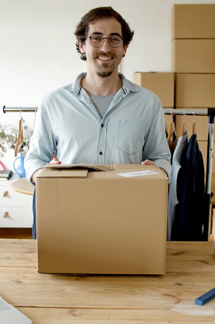 A man with glasses and dark curly hair, wearing a light blue button-down shirt, is smiling while holding a medium-sized cardboard box sealed with packing tape, positioned on a wooden table. The background features a domestic setting with a clothing rail holding jackets and shirts, a window with natural light, and several cardboard boxes stacked on a shelf. The scene depicts packing activities related to house removals, integral to home relocation and furniture transport services provided by Man and a Van Catford. The lighting is natural and even, highlighting the packing process and indicating preparation for a house move or small business relocation within the context of professional removal services.