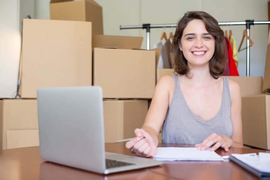A male professional mover with a dark beard and curly hair, smiling and standing behind a stack of cardboard boxes in a well-lit room with a large arched window in the background. The boxes are of various sizes, some sealed with red packing tape, and are likely used for packing office furniture, equipment, or personal belongings as part of a house or office relocation service. The movers are inside a residential or commercial property during the packing and moving process, with the boxes placed on a clean floor near the doorway. The natural lighting coming through the window illuminates the space, highlighting the orderly arrangement of packing materials and the environment prepared for furniture transport. This scene exemplifies efficient packing and loading activities conducted by [COMPANY_NAME], engaged in house or office removals, ensuring organized and professional moving logistics.