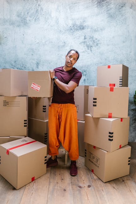 Two individuals engaged in a home relocation process, with one person seated inside a property and the other kneeling beside a large cardboard box filled with various items. The person inside the prop