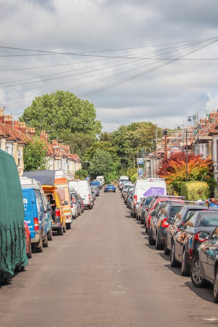 A narrow residential street lined with parked cars on both sides, including a variety of vehicles such as vans, small commercial trucks, and passenger cars, some partially covered with tarpaulins or protective sheets. The pavement is asphalt, with houses and terraced homes visible along the street, featuring brick facades, front gardens, and small trees or shrubs. Overhead, power lines stretch across the street, and the sky above is partly cloudy with patches of blue. This scene captures the typical environment encountered during a home relocation in Catford, highlighting the tight street access that requires careful planning for furniture transport and loading for a house removal. The presence of multiple parked vehicles indicates the need for precise organisation during the loading process by professional movers like Man and a Van Catford, who often deal with similar logistics in narrow street access situations.