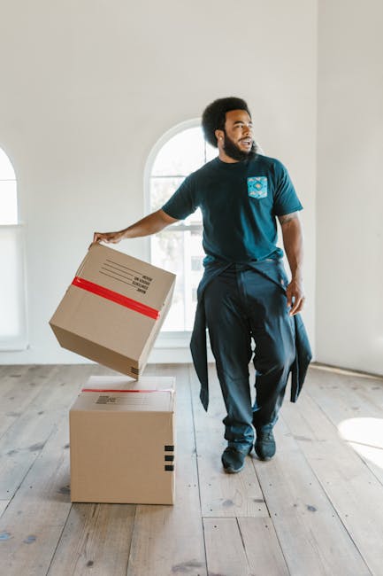 A man with a dark beard and curly black hair, wearing a dark blue t-shirt with a blue logo on the chest and dark trousers, is inside a well-lit room with light-colored walls and wooden flooring. He is holding a cardboard box with red tape around it, preparing to move it, as part of a home relocation process. Behind him, a large arched window allows natural light to enter, illuminating his face and the surrounding area. On the floor in front of him, there are two additional cardboard boxes, one stacked on top of the other, both sealed with packing tape. The scene captures a moment of furniture transport and packing during the house removal, with no other furniture or objects visible in the room. This image illustrates the careful handling and organization involved in professional removals, such as those provided by Man and a Van Catford, supporting efficient moving and loading processes.